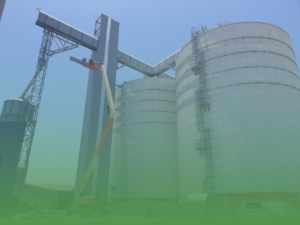 Worker in an elevated lift performing maintenance on tall industrial storage silos and conveyor infrastructure.